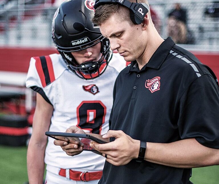 American-Football-Coach analysiert mit einem jungen Spieler Spielzüge auf einem Tablet am Spielfeldrand.